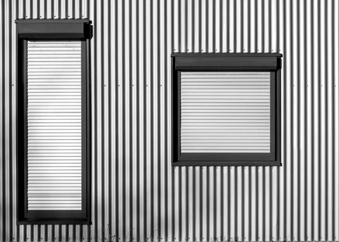 Dwa okna aluminiowe z roletami zewnętrznymi w budynku o elewacji z blachy falistej.